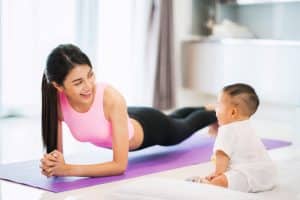 Woman smiles on yoga mat with a restored physique beside her toddler.